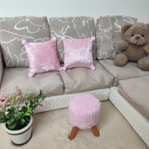 K&ouml;nnte beinhalten: Ein wei&szlig;es Sofa mit drei beigen Kissen und zwei rosa Samtkissen. Ein brauner Teddyb&auml;r sitzt auf dem Sofa. Ein rosa, flauschiger Hocker mit Holzbeinen steht vor dem Sofa. Eine kleine Topfpflanze steht auf dem Boden vor dem Sofa.