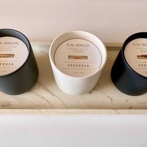 May include: Three candles in black, white, and black containers. The candles are labeled "SUN-ROW.CO." and "PODEROSA". The candles are on a light gray concrete tray.