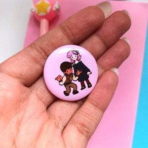 May include: A pink button featuring a cartoon illustration of two people. The person on the left is wearing a brown shirt and trousers and has brown hair. The person on the right is wearing a purple shirt and black skirt and has purple hair.