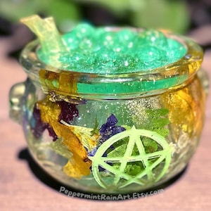 Peut inclure: Un chaudron en verre transparent rempli d'une matière verte et scintillante et de fleurs séchées. Le chaudron porte un symbole de pentacle vert néon à l'avant et une poignée de chaque côté. Le texte "PeppermintRainArt.Etsy.com" est en bas.