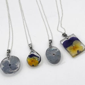 Puede incluir: Cuatro collares plateados con colgantes que contienen flores prensadas. Los colgantes tienen varias formas, incluyendo redonda, de lágrima y rectangular. Las flores son pensamientos amarillos y morados encapsulados en resina transparente.