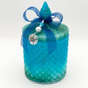 以下が含まれることがあります： ダイヤモンド模様のターコイズガラスジャー。お揃いの蓋と、リボンで結ばれた透明な青いリボンが付いています。砂ドルデザインのシルバーチャームがリボンからぶら下がっています。ジャーは、水色と緑色のグラデーションカラーです。
