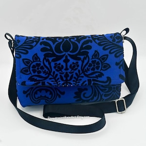 May include: A blue crossbody bag with a black floral pattern. The bag has a black adjustable strap and a silver-colored buckle. The bag's flap is closed, and the text "PeppermintRainArt.Etsy.com" is visible on the bottom.