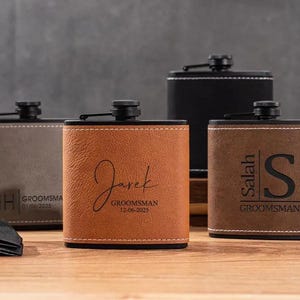 May include: Personalised hip flasks in brown, grey, and black. Each flask has custom text, including names and dates, with a black cap and a leather-like texture. The flasks are displayed on a wooden surface, with a bow tie in the foreground.