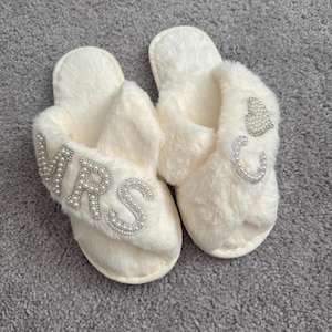 May include: A pair of cream-coloured, fluffy slippers. One slipper is embroidered with the word "MRS" in pearl-like beads. The other slipper has a pearl-like heart and the letter "C". The slippers are on a grey surface.