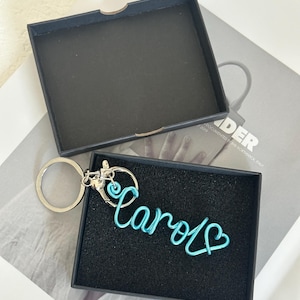 May include: A teal wire keychain spelling "Carol" in cursive, with a heart detail at the end. The keychain is presented in an open black gift box with a black interior. The background is a light-coloured surface.