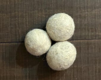 Puede incluir: Tres bolas de secadora de lana gris claro dispuestas sobre una superficie de madera marrón oscuro. Las bolas son redondas y tienen un aspecto texturizado y fibroso. El fondo es una veta de madera oscura.