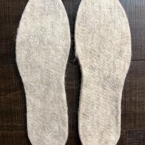 May include: Two light gray wool felt insoles, designed to fit inside shoes. The insoles are oval-shaped, with a textured surface. The insoles are placed side by side on a dark brown wooden surface.