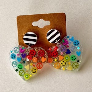 Puede incluir: Pendientes en forma de coraz&oacute;n con una base transparente y brillante. Los pendientes est&aacute;n adornados con frutas coloridas, caras sonrientes, s&iacute;mbolos de la paz y estrellas. Los tachuelas son c&iacute;rculos a rayas blancas y negras.