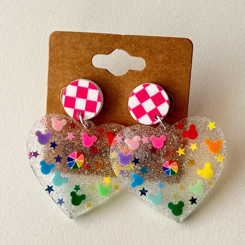 Puede incluir: Pendientes transparentes en forma de coraz&oacute;n con siluetas coloridas de Mickey Mouse, estrellas y un dise&ntilde;o de arco&iacute;ris. Los pendientes tienen una base de purpurina plateada y est&aacute;n rematados con un c&iacute;rculo a cuadros rosa y blanco. Los pendientes est&aacute;n sujetos a un respaldo de cart&oacute;n marr&oacute;n.