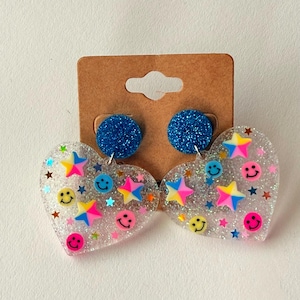 Puede incluir: Pendientes transparentes en forma de coraz&oacute;n con purpurina, caras sonrientes y estrellas de colores. Los pendientes tienen tachuelas azules con purpurina.