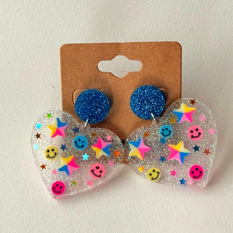 Puede incluir: Pendientes transparentes en forma de coraz&oacute;n con purpurina, caras sonrientes y estrellas de colores. Los pendientes tienen tachuelas azules con purpurina.