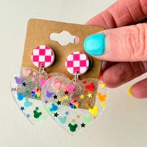 Puede incluir: Pendientes transparentes en forma de coraz&oacute;n con siluetas coloridas de Mickey Mouse, estrellas y detalles de arco&iacute;ris. Los pendientes tienen un c&iacute;rculo con estampado de cuadros rosas y blancos. Presentados en cart&oacute;n marr&oacute;n.