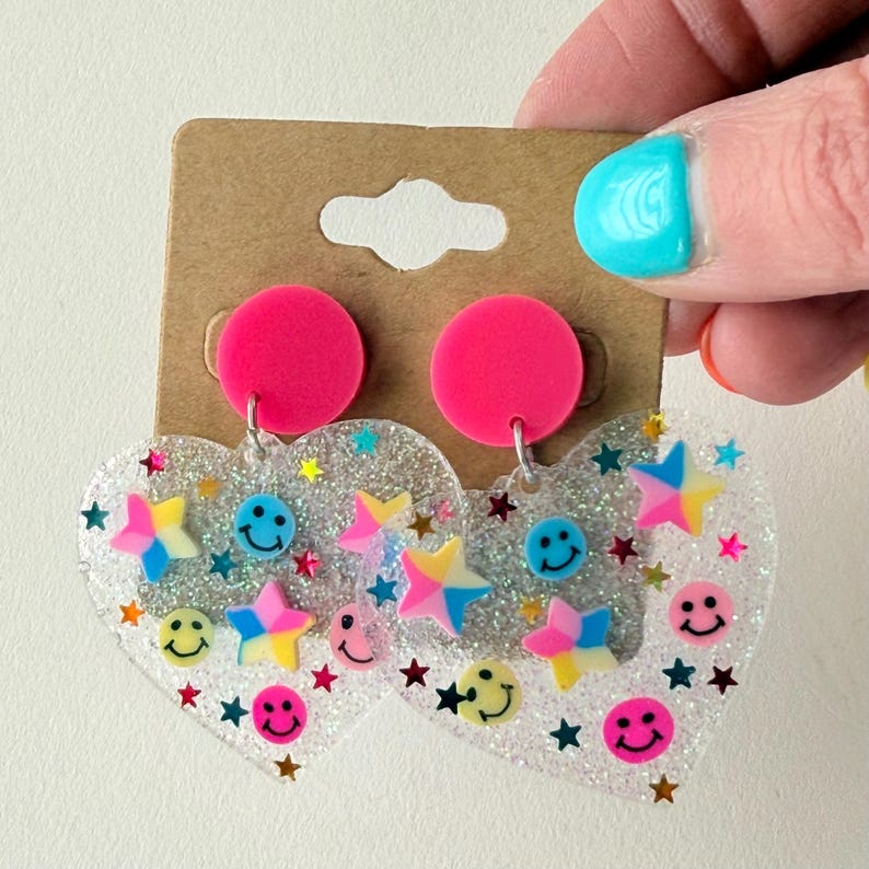 Puede incluir: Un par de pendientes en forma de coraz&oacute;n con un dise&ntilde;o transparente y brillante. Los pendientes presentan estrellas de colores, caras sonrientes y tachuelas circulares rosas. Los pendientes se sostienen contra una etiqueta marr&oacute;n.