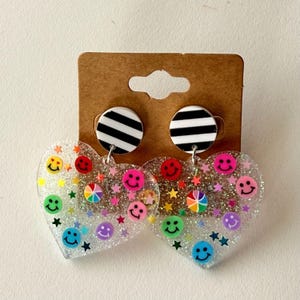 Puede incluir: Pendientes en forma de coraz&oacute;n con base transparente y brillante. Presentan caras sonrientes de colores y estrellas. La parte superior de cada pendiente tiene un c&iacute;rculo a rayas blancas y negras.