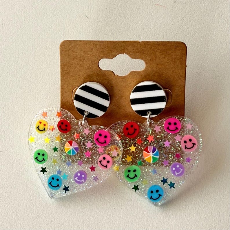 Puede incluir: Pendientes en forma de coraz&oacute;n con base transparente y brillante. Presentan caras sonrientes de colores y estrellas. La parte superior de cada pendiente tiene un c&iacute;rculo a rayas blancas y negras.