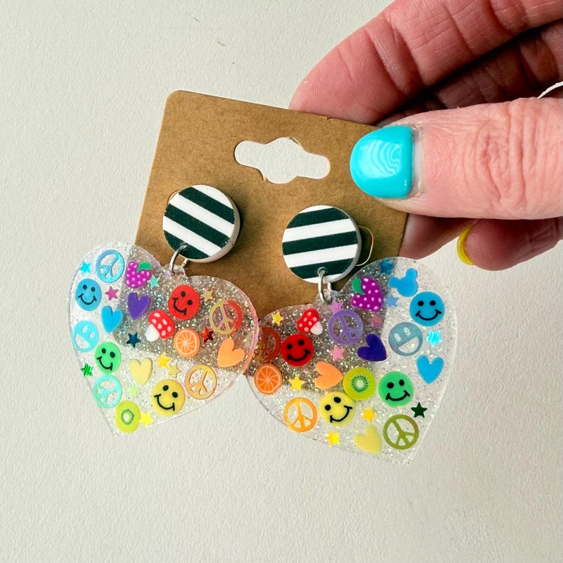 Puede incluir: Pendientes transparentes en forma de coraz&oacute;n con caras sonrientes de colores, s&iacute;mbolos de la paz y dise&ntilde;os de frutas. Los pendientes tienen un aspecto brillante y est&aacute;n sujetos a tachuelas circulares a rayas blancas y negras. Los pendientes est&aacute;n sostenidos por una mano.
