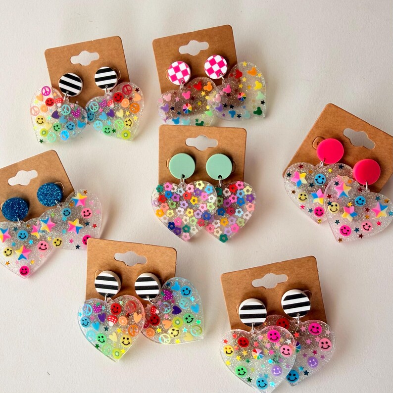 Puede incluir: Una colecci&oacute;n de pendientes en forma de coraz&oacute;n con varios dise&ntilde;os. Algunos pendientes presentan caras sonrientes, estrellas y s&iacute;mbolos de la paz, mientras que otros tienen dise&ntilde;os florales. Los pendientes est&aacute;n en tarjetas de cart&oacute;n marr&oacute;n.