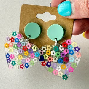 Puede incluir: Pendientes transparentes en forma de coraz&oacute;n llenos de confeti con forma de flor de colores. Los pendientes tienen tachuelas verde menta y ganchos plateados. Los pendientes est&aacute;n sujetos a una etiqueta de cart&oacute;n marr&oacute;n.