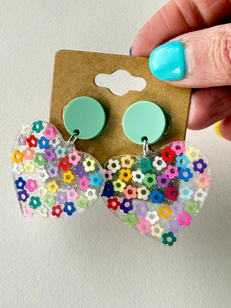 Puede incluir: Pendientes transparentes en forma de coraz&oacute;n llenos de confeti con forma de flor de colores. Los pendientes tienen tachuelas verde menta y ganchos plateados. Los pendientes est&aacute;n sujetos a una etiqueta de cart&oacute;n marr&oacute;n.
