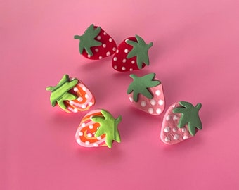 May include: Six strawberry-shaped decorative objects with green leaves and white dots on a pink background. The strawberries are red, pink, and orange. The objects are likely made of clay or a similar material.