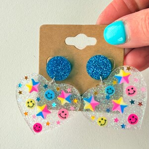 Puede incluir: Un par de pendientes en forma de coraz&oacute;n con un acabado transparente y brillante. Los pendientes presentan estrellas de colores y caras sonrientes. La parte superior de los pendientes son c&iacute;rculos de purpurina azul.
