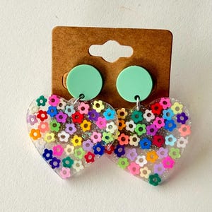 Puede incluir: Pendientes transparentes en forma de coraz&oacute;n llenos de flores de colores. Los pendientes tienen un c&iacute;rculo verde menta en la parte superior y est&aacute;n sujetos a una tarjeta marr&oacute;n.