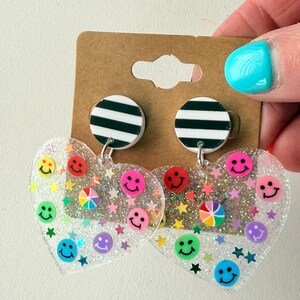 Puede incluir: Un par de pendientes transparentes en forma de coraz&oacute;n con caras sonrientes de colores y confeti en forma de estrella. Los pendientes tienen una parte superior circular a rayas en blanco y negro. Los pendientes est&aacute;n sujetos a un respaldo de cart&oacute;n marr&oacute;n.
