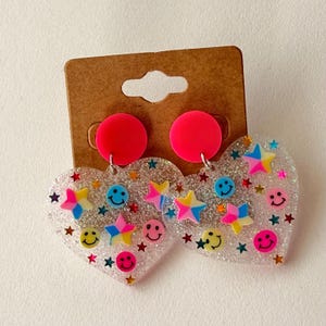 Puede incluir: Pendientes en forma de coraz&oacute;n con base transparente y brillante. Presentan dise&ntilde;os de estrellas y caras sonrientes de colores. Cada pendiente tiene un perno circular rosa brillante.