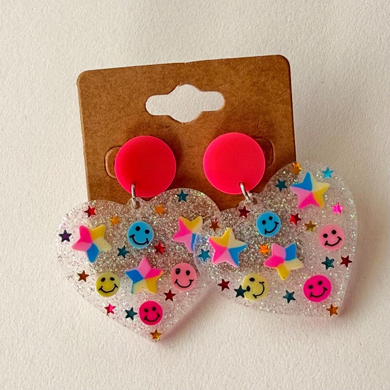 Puede incluir: Pendientes en forma de coraz&oacute;n con base transparente y brillante. Presentan dise&ntilde;os de estrellas y caras sonrientes de colores. Cada pendiente tiene un perno circular rosa brillante.
