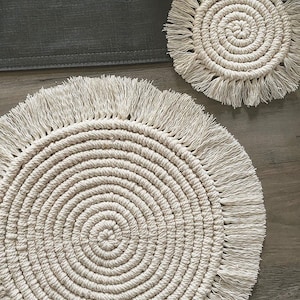 May include: Two round, white, crocheted coasters with fringe. The coasters are made of a thick, rope-like material and have a circular pattern.