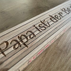 Pode incluir: Uma régua dobrável de madeira com as palavras "Papa é o melhor" gravadas na parte superior. A régua tem um fundo branco com marcas vermelhas.