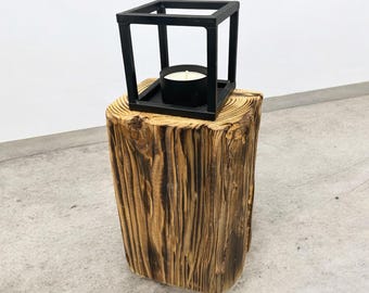 May include: A rustic, wooden side table with a natural, textured finish. A black metal lantern with a candle inside sits on top. The lantern has a square frame design, creating a modern contrast to the organic wood.