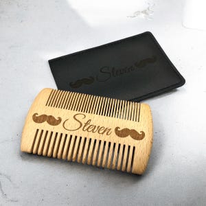 May include: A wooden beard comb with engraved mustache designs and the name "Steven". The comb is light brown and has closely spaced teeth. A black leather case with the name "Steven" and a mustache design is behind the comb.