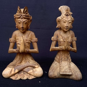 Puede incluir: Dos estatuas de madera de personas en posición sentada con las manos juntas en oración. Las estatuas están talladas en un estilo tradicional y tienen detalles intrincados. Las estatuas están hechas de madera marrón claro.