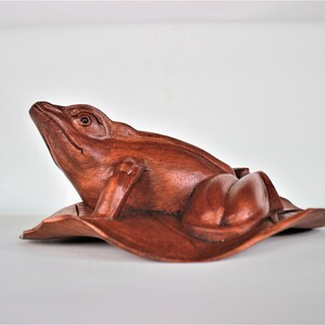 May include: A hand-carved wooden frog figurine, painted in a rich reddish-brown color, is perched on a leaf. The frog is looking up with its mouth slightly open, and its eyes are detailed with black pupils.