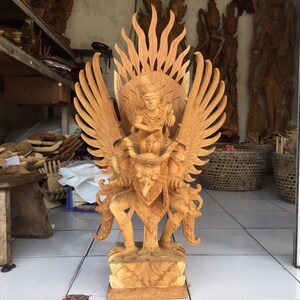 May include: A wooden sculpture of a mythical bird-like creature with outstretched wings. The creature is holding a human figure in its talons. The sculpture is carved in a detailed and intricate style, with a natural wood finish.