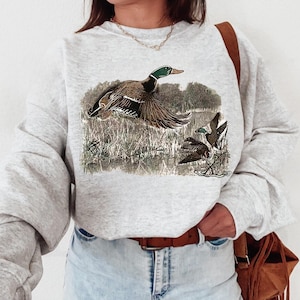 May include: A gray sweatshirt with a vintage illustration of a duck flying over a marsh. The duck is brown and green with a white breast. The sweatshirt has long sleeves and a crew neck.