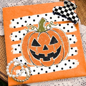 May include: A square orange sign featuring a central jack-o'-lantern with a green stem and black facial features. The pumpkin is set against a white background with black polka dots. A black and white checkered hair claw and a gold beaded bracelet.