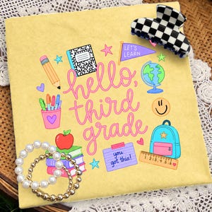 Pode incluir: Uma t-shirt amarela com um gráfico de temática escolar com o texto "hello, third grade" e "you got this!" rodeado por ilustrações de um lápis, caderno, globo, mochila, régua, livros e uma maçã, com uma mola de cabelo axadrezada e pulseiras de pérolas.