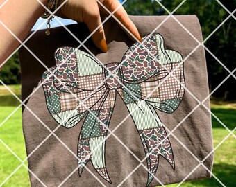 May include: A brown t-shirt featuring a large bow design. The bow is composed of various patterned patches in shades of green, brown, and tan. The shirt is held up against a background of green grass and a blue sky.