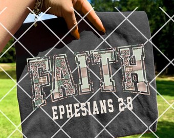 Könnte beinhalten: Dunkelgraues T-Shirt mit dem Wort "FAITH" in einem Schriftstil im College-Stil, mit einem Tarnmuster in den Buchstaben. Der Text "EPHESIANS 2:8" ist darunter in Weiß gedruckt. Das T-Shirt wird vor dem Hintergrund von grünem Gras und blauem Himmel gehalten.