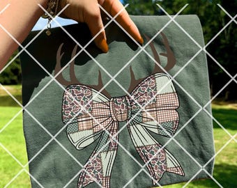 May include: Olive green t-shirt featuring a large bow design with a patchwork pattern in shades of brown, green, and white. The bow is adorned with brown antlers. The shirt is held up against a backdrop of green grass and a blue sky.