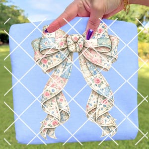 May include: A light blue square with a large bow illustration. The bow features a patchwork design with pink roses and blue accents. The bow is held by a hand with purple nail polish.