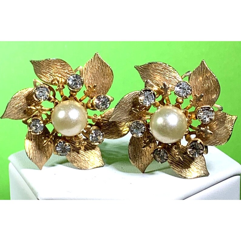 Puede incluir: Un par de pendientes de oro en forma de flor con una perla y diamantes de imitaci&oacute;n en el centro de cada flor.