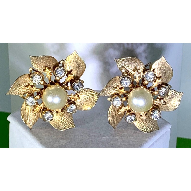 Puede incluir: Un par de pendientes de tono dorado con dise&ntilde;o de flor. Cada pendiente presenta una perla falsa en el centro rodeada de diamantes de imitaci&oacute;n transparentes.