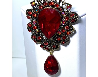 Pode incluir: Um broche dourado com um grande cristal vermelho em forma de lágrima no centro. O broche é rodeado por cristais vermelhos menores e tem um cristal vermelho menor em forma de lágrima pendurado na parte inferior.
