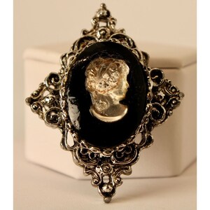 Puede incluir: Un broche de camafeo plateado con fondo negro y un camafeo blanco con el perfil de una mujer.