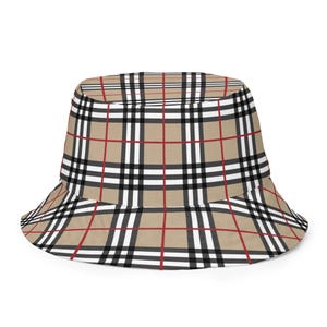 Op de afbeelding: Een beige vissershoed met een ruitpatroon in zwart, wit en rood. De hoed heeft een brede rand en een klassieke, casual stijl. Deze hoed is een modieus accessoire.
