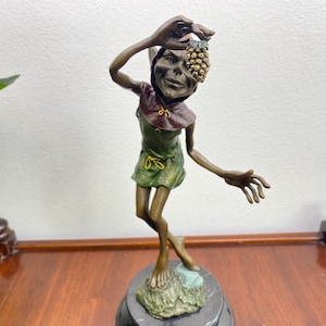 May include: A bronze-toned figurine of a pointed-eared creature wearing a green tunic and a dark hood. The figure stands on a textured base, holding a small, golden, textured cluster of fruit above its head with one hand while gesturing outward.
